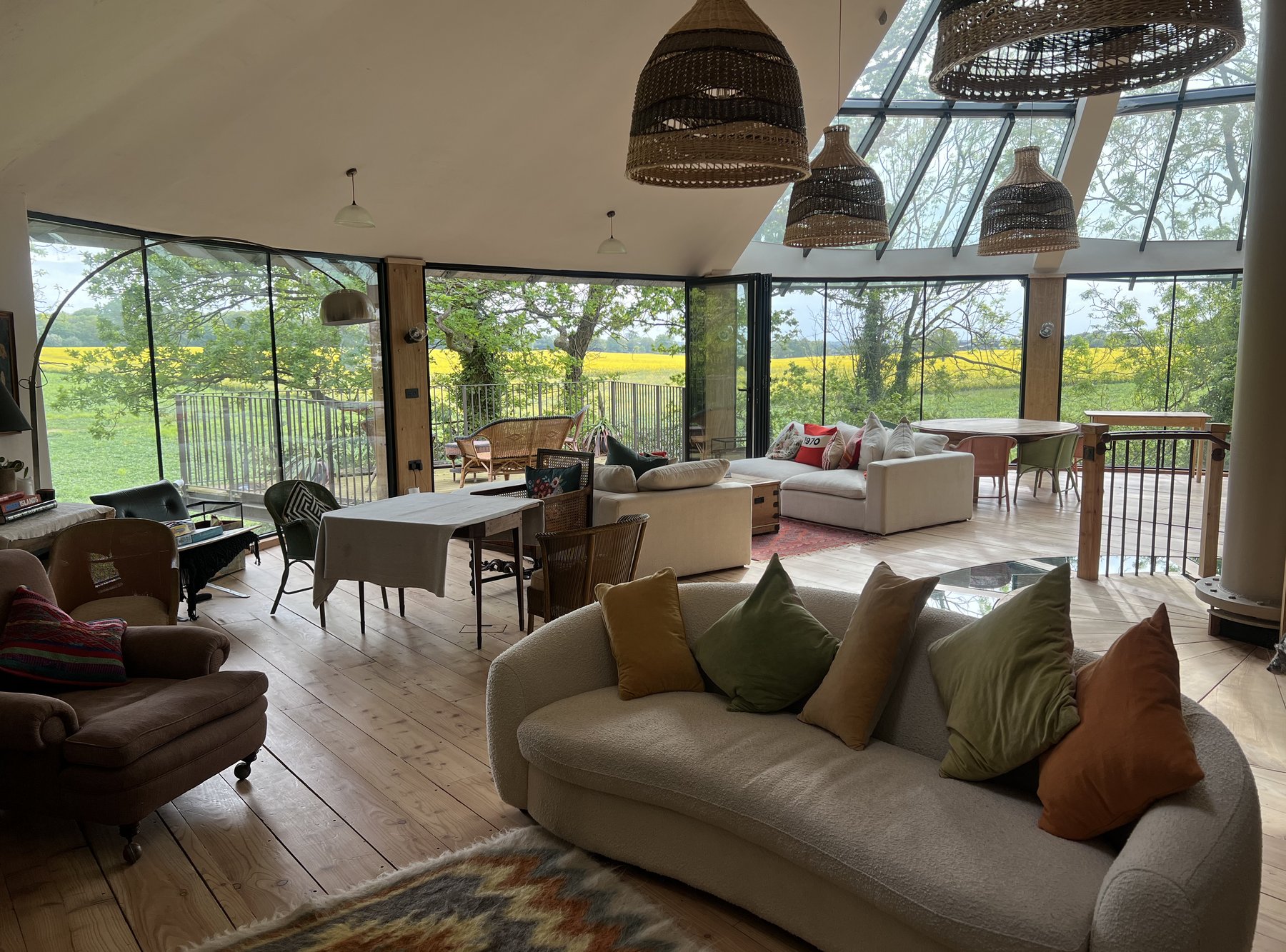 Wide view of the open-plan top floor — pendants, curved sofa, dining and floor-to-ceiling Crittall glass looking out across the field